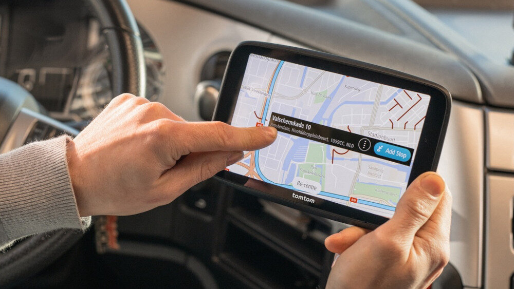 Osoba trzyma urządzenie nawigacyjne z wyświetloną mapą i trasą, na ekranie widoczny jest tekst: \'Valschermkade 10\'. Na dole ekranu znajduje się logo \'tomtom\'.