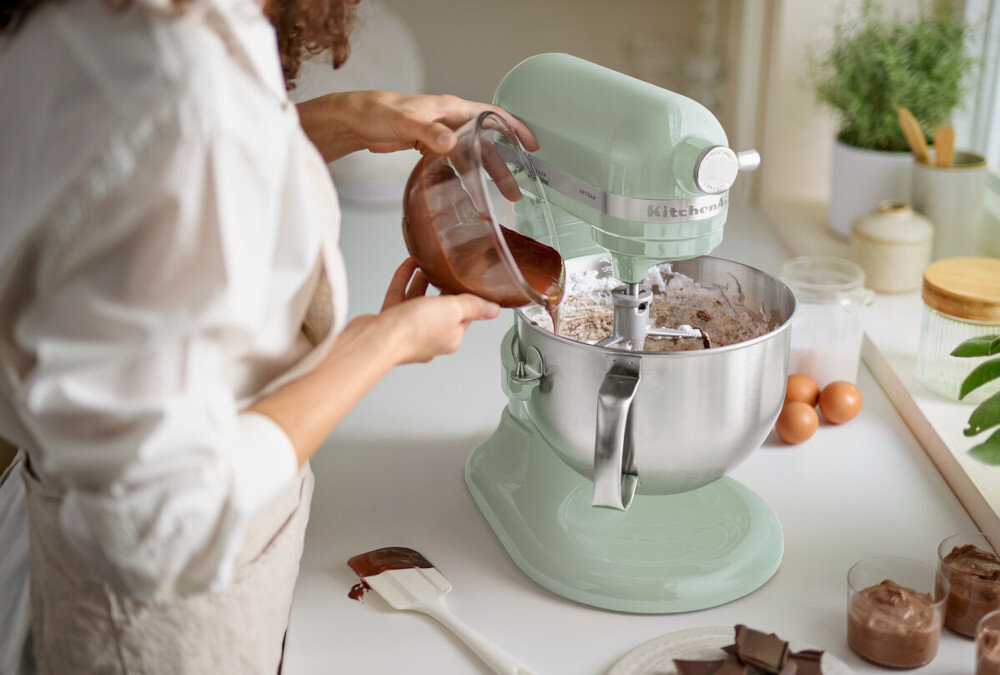 Osoba wlewa płyn do misy robota kuchennego w kolorze pistacjowym z widocznym logo \'KitchenAid\'. Na blacie znajdują się jajka, czekolada i inne składniki do pieczenia.