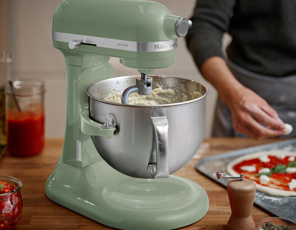 Pistacjowy robot kuchenny planetarny z misą ze stali nierdzewnej, w tle osoba przygotowująca pizzę. Na blacie widoczne są słoiki z sosem i przyprawami oraz młynek do pieprzu.