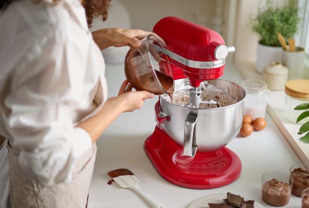 Osoba wlewa płyn z miski do czerwonego robota kuchennego marki KitchenAid, który stoi na blacie kuchennym. W tle widoczne są jajka, rośliny i inne akcesoria kuchenne.