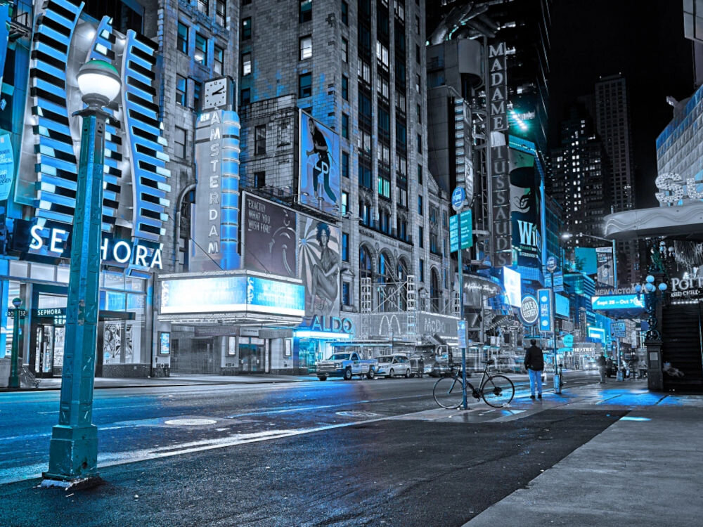 Nocna scena miejska z neonowymi reklamami, w tym widoczne napisy \'SEPHORA\', \'ALDO\', \'Amsterdam\' i \'Madame Tussauds\'. Na ulicy stoi kilka samochodów, a w tle widać wysokie budynki.
