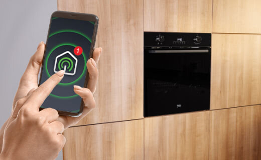 Osoba trzyma smartfon z aplikacją pokazującą ikonę domu z powiadomieniem. Obok znajduje się czarny piekarnik w zabudowie z widocznym logo Beko.
