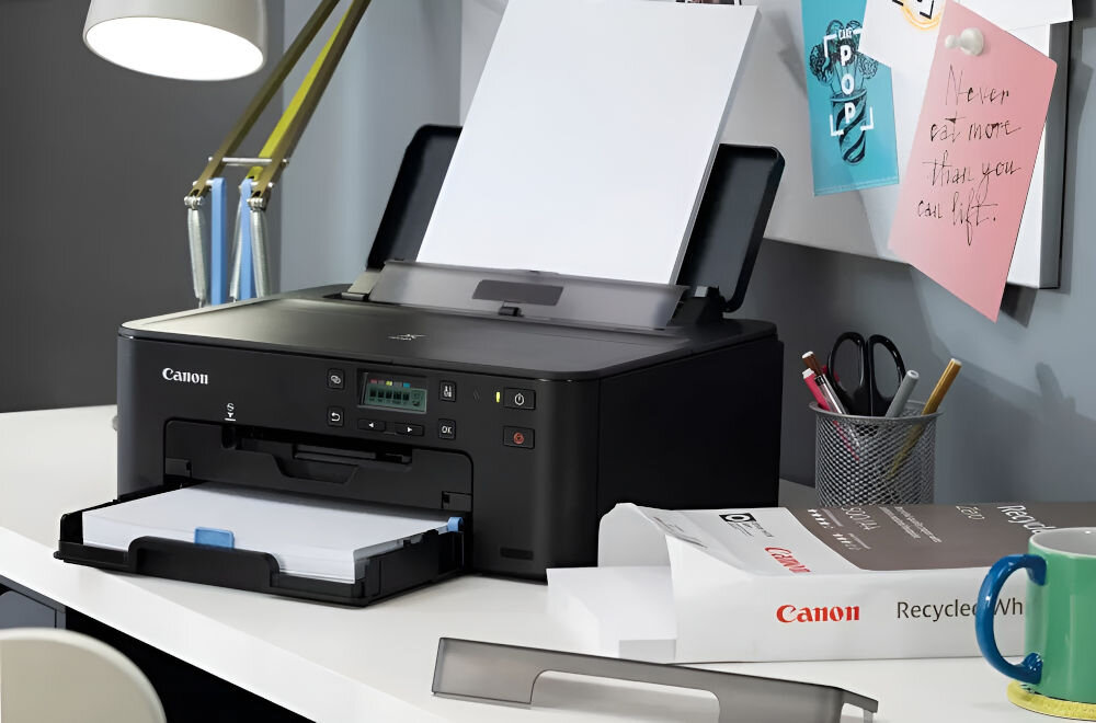 Na biurku znajduje się czarna drukarka Canon z otwartą tacą na papier. Obok leży pudełko z napisem \'Canon Recycled White\'.