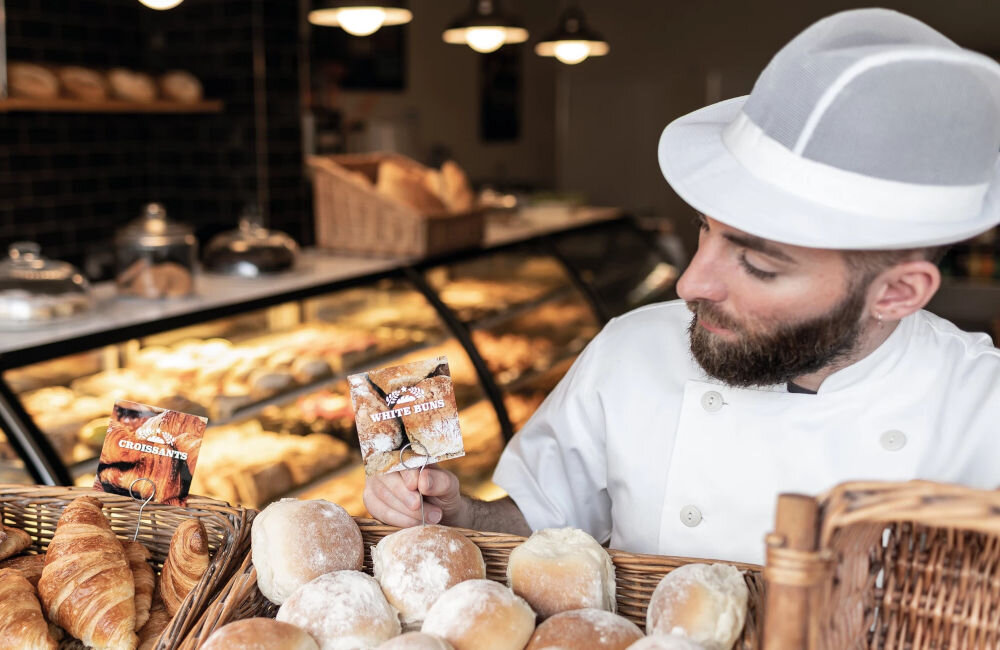 Mężczyzna w białym stroju piekarza trzyma koszyk z bułkami, obok którego znajdują się etykiety z napisami \'CROISSANTS\' i \'WHITE BUNS\'. W tle widać ladę z różnymi wypiekami.