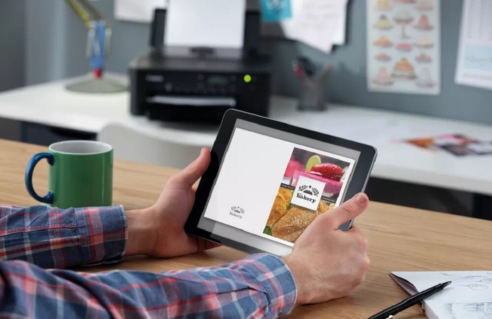 Osoba trzyma tablet z wyświetlonym obrazem przedstawiającym produkty piekarnicze i napis \'Bakery\'. W tle znajduje się drukarka na biurku oraz kubek.