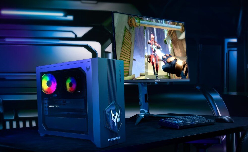 Na obrazie znajduje się komputer stacjonarny z podświetleniem RGB oraz monitor wyświetlający grę wideo. Obok komputera widoczna jest klawiatura i myszka, a na obudowie komputera znajduje się logo producenta.