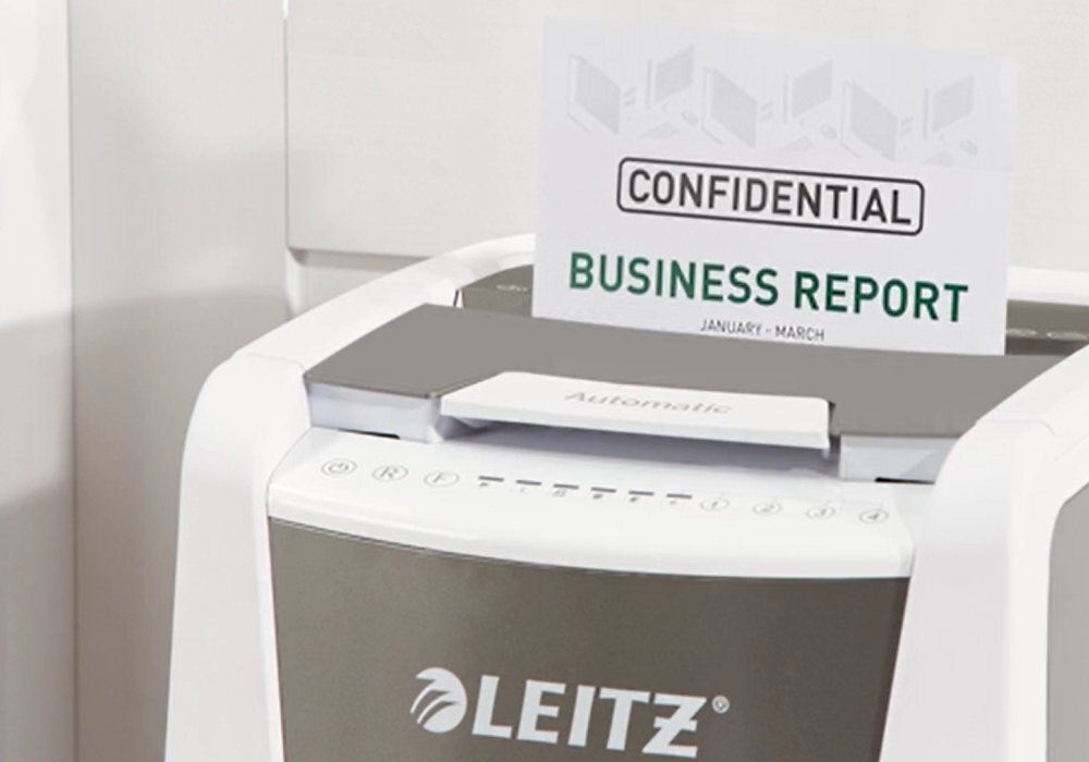 Na obrazie widoczna jest niszczarka z logo \'LEITZ\', do której wkładany jest dokument z napisem \'CONFIDENTIAL BUSINESS REPORT JANUARY - MARCH\'.