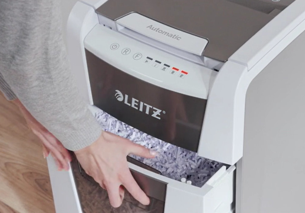 Osoba otwiera szufladę niszczarki z widocznymi pociętymi papierami. Na urządzeniu widoczne jest logo \'LEITZ\' oraz napis \'Automatic\'.