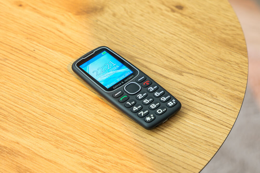 Na drewnianym stole leży czarny telefon komórkowy z klawiaturą numeryczną. Na ekranie widoczny jest czas 22:21 oraz napis \'Götze & Jensen\'.