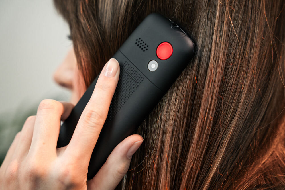 Osoba trzyma przy uchu czarny telefon z czerwonym przyciskiem i kratką głośnika. Telefon ma matową powierzchnię i widoczne są detale konstrukcyjne.