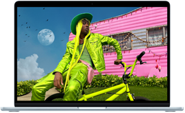 Na ekranie laptopa widać osobę w zielonym stroju siedzącą na rowerze przed różowym budynkiem. W tle znajduje się księżyc i niebo z chmurami.