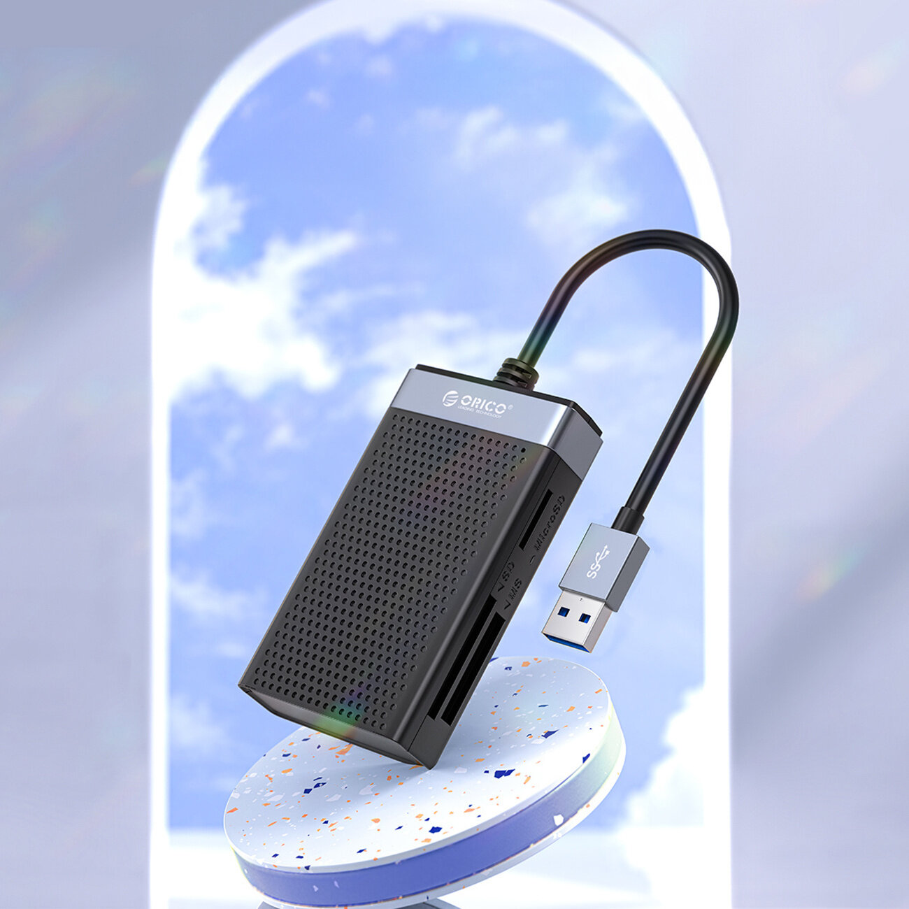 Na obrazie widoczny jest czarno-srebrny czytnik kart pamięci z logo \'Orico\' oraz podłączonym kablem USB. W tle znajduje się niebieskie niebo z białymi chmurami widoczne przez łukowate okno.