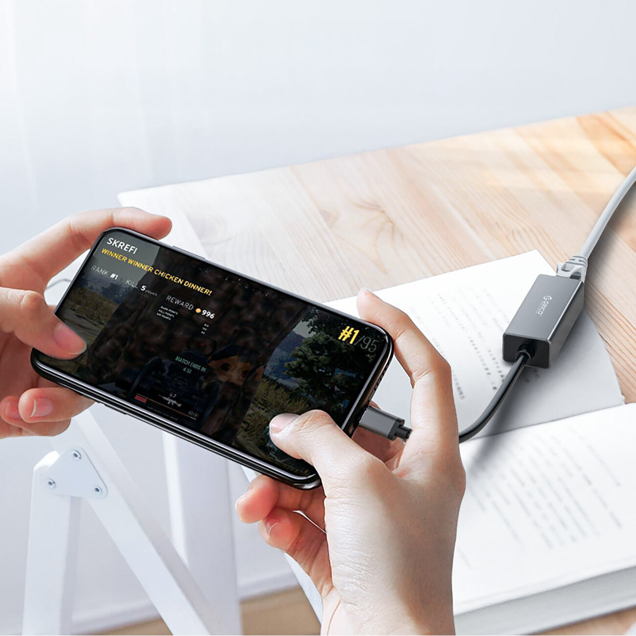 Osoba trzyma smartfon z wyświetlonym ekranem gry, podłączony do adaptera sieciowego z logo Orico, który leży na drewnianym blacie obok otwartej książki.
