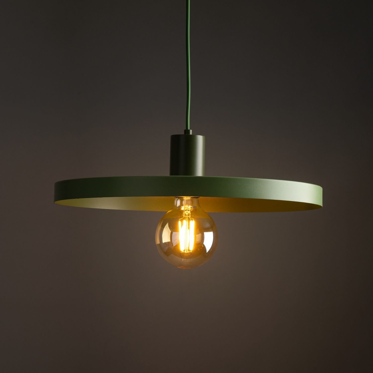 Zielona lampa wisząca z okrągłym kloszem i widoczną żarówką, zawieszona na ciemnym tle.