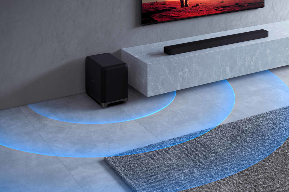 Na obrazie widać czarny soundbar umieszczony na szarej, marmurowej półce oraz subwoofer stojący na podłodze. Na podłodze widoczne są niebieskie, półkoliste linie sugerujące rozchodzenie się dźwięku.