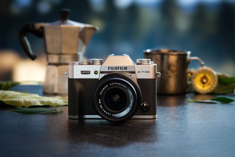 Na zdjęciu znajduje się aparat fotograficzny z logo \'FUJIFILM X-T30\' na obudowie, ustawiony na ciemnym blacie. W tle widoczne są metalowy dzbanek i kubek oraz liście.