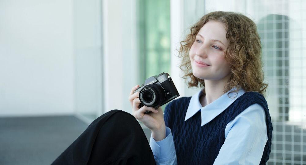 Osoba trzyma aparat fotograficzny, siedząc w jasnym pomieszczeniu. Aparat jest czarny z widocznym obiektywem.
