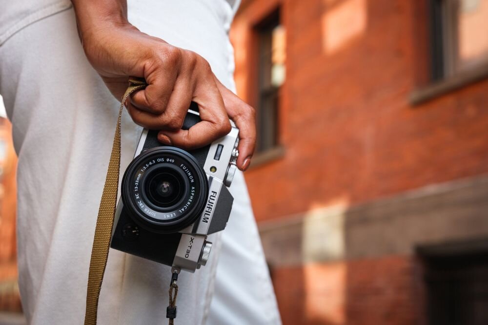 Osoba trzyma aparat fotograficzny z widocznym logo \'Fujifilm\' i oznaczeniem modelu \'X-T30\'. W tle ceglana ściana budynku.