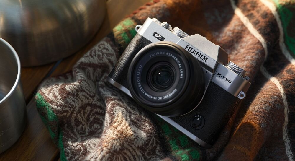 Aparat fotograficzny z logo \'FUJIFILM\' i oznaczeniem modelu \'X-T30\' leży na wzorzystym materiale. Obiektyw aparatu ma widoczny napis \'FUJINON ASPHERICAL LENS\'.