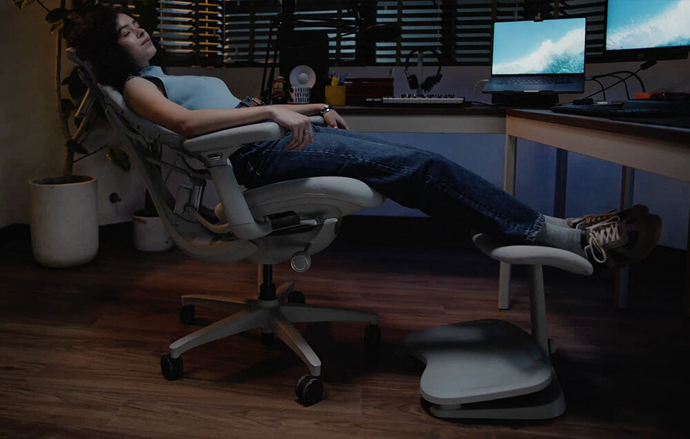 Osoba siedzi na ergonomicznym krześle, opierając nogi na podnóżku. W tle znajduje się biurko z dwoma monitorami i różnymi przedmiotami biurowymi.