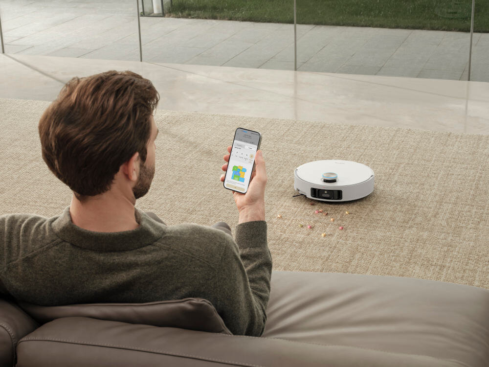 Osoba siedzi na kanapie, trzymając w ręku smartfon z aplikacją pokazującą mapę. Na dywanie obok znajduje się biały robot sprzątający, wokół którego leżą rozsypane drobne przedmioty.