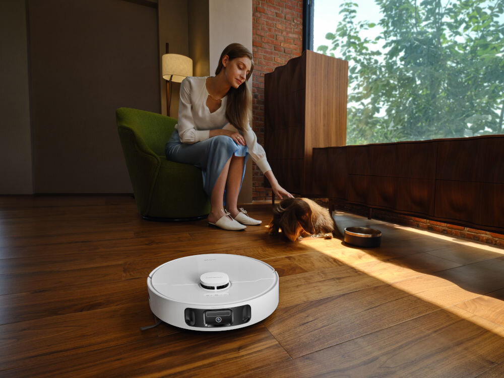 Na drewnianej podłodze znajduje się biały robot sprzątający. W tle kobieta siedzi na fotelu i głaszcze psa.