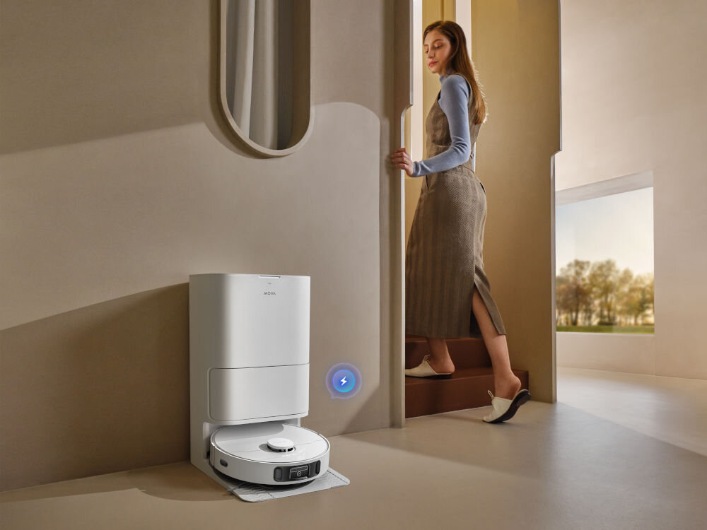 Na obrazie widoczny jest robot sprzątający MOVA E40 Ultra w stacji dokującej, umieszczony przy ścianie. W tle znajduje się osoba wchodząca po schodach oraz duże okno z widokiem na zewnątrz.