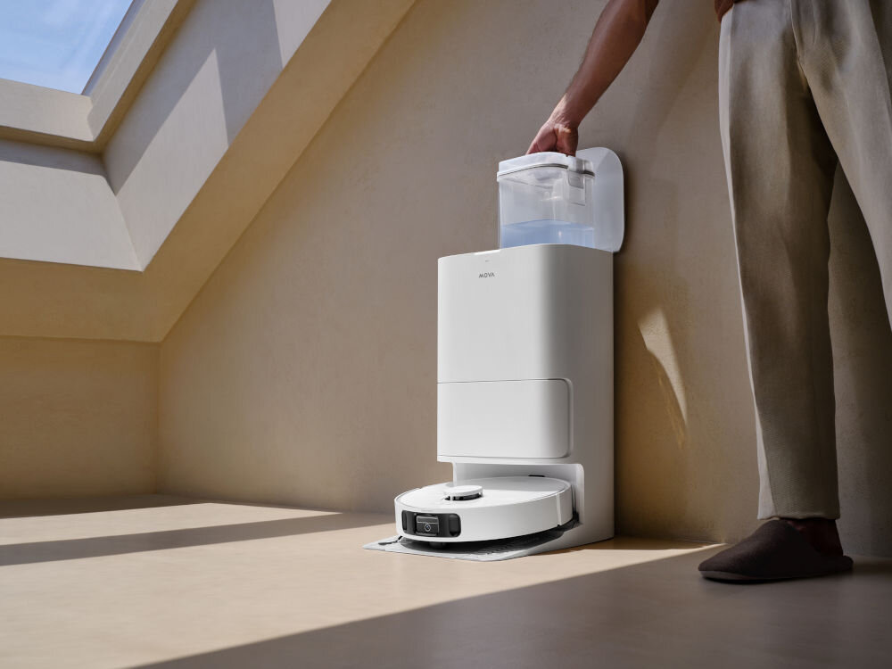 Na podłodze znajduje się robot sprzątający w stacji dokującej, obok której stoi osoba trzymająca pojemnik z wodą. Na stacji widoczne jest logo \'MOVA\'.