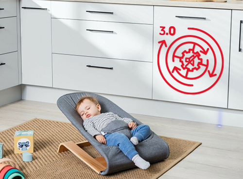 Dziecko śpi w foteliku na podłodze w kuchni, obok szafek. Na szafce widoczny jest czerwony symbol zębatki z napisem \'3.0\'.
