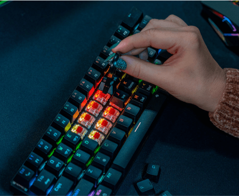 Osoba trzyma klawisz nad klawiaturą z podświetleniem RGB, na której widać kilka zdjętych klawiszy. Klawiatura jest czarna, a podświetlenie ma różne kolory.