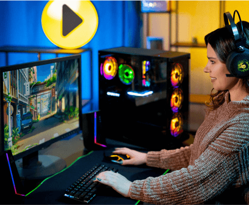Osoba siedzi przy biurku, używając klawiatury i myszy, patrząc na monitor z grą. W tle widać komputer z podświetleniem RGB oraz duże logo w kształcie żółtego przycisku odtwarzania.