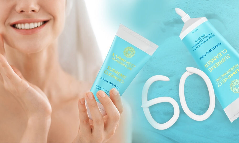 Na obrazie widoczna jest osoba trzymająca tubkę z napisem \'LUMINEO SUPREME CLEANSER FOR ALL SKIN TYPES\'. Obok znajduje się druga tubka z tym samym napisem oraz liczba \'60\' ułożona z kremu na niebieskim tle.
