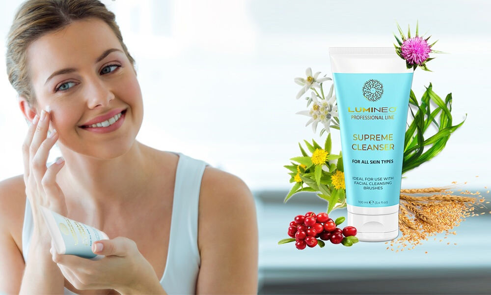 Kobieta uśmiecha się, trzymając tubkę z napisem \'LUMINEO PROFESSIONAL LINE SUPREME CLEANSER FOR ALL SKIN TYPES\'. Obok widoczna jest większa tubka żelu otoczona kwiatami i owocami.