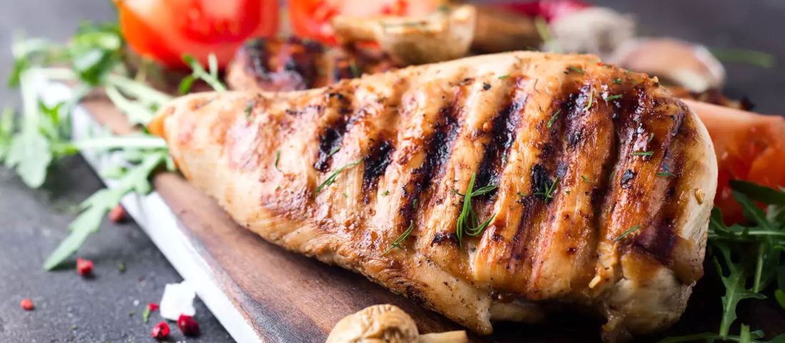 Na desce leży grillowana pierś z kurczaka z widocznymi śladami grillowania, obok znajdują się pomidory i rukola.