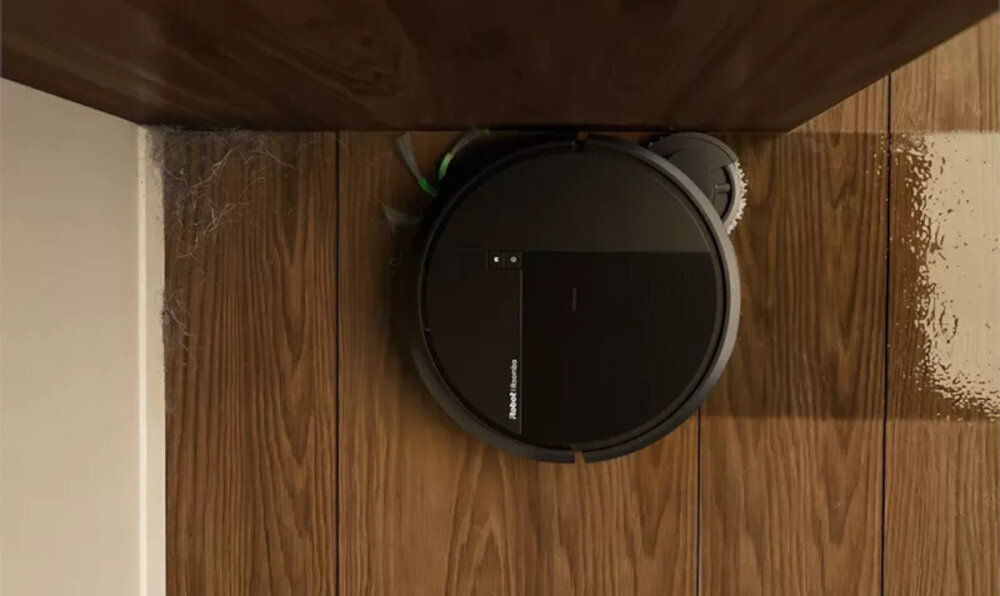 Czarny robot sprzątający znajduje się na drewnianej podłodze przy ścianie, z widocznym logo \'Roomba\'.