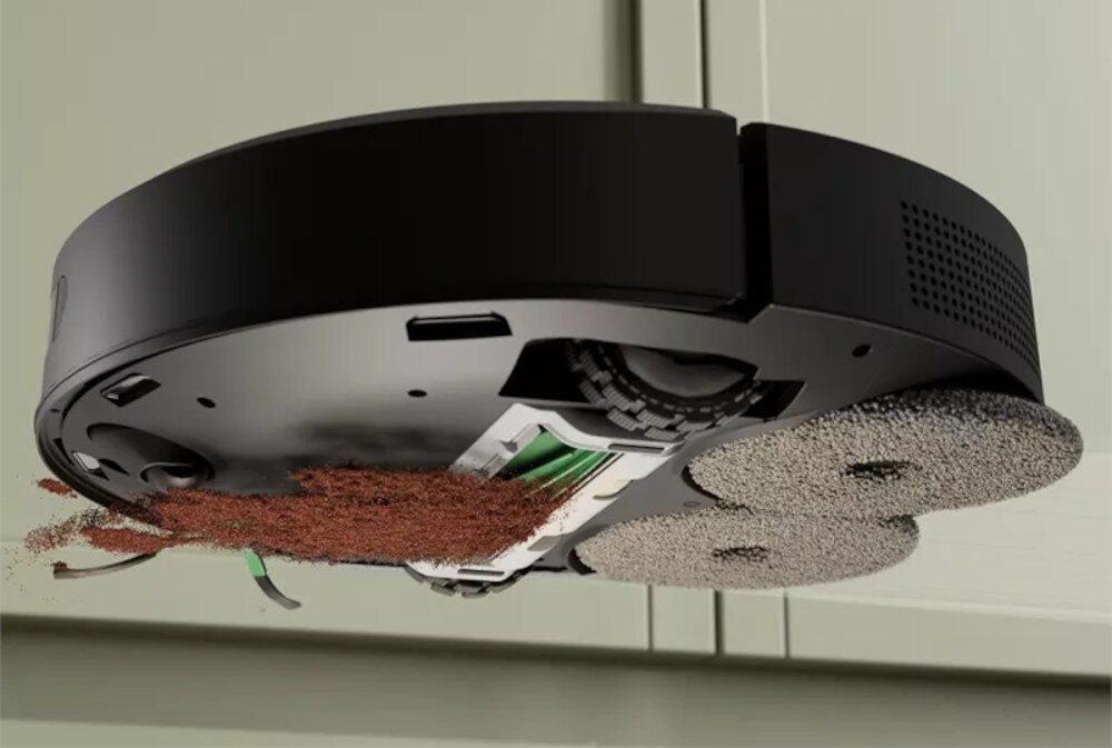 Czarny robot sprzątający z widocznymi szczotkami i otworem zasysającym, z którego wysypuje się brązowy materiał. Urządzenie znajduje się na tle jasnej powierzchni.