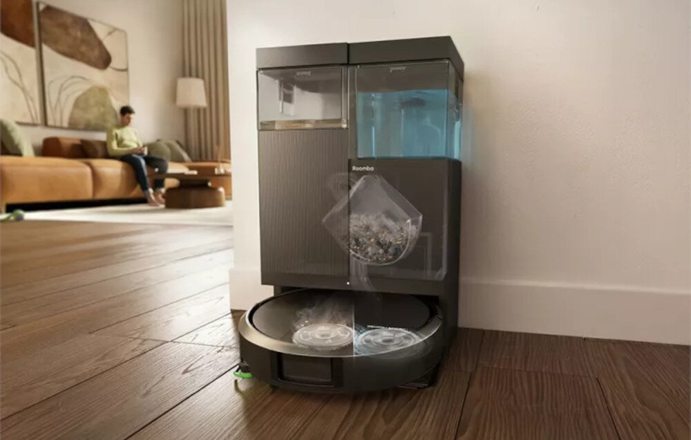 Na podłodze w pomieszczeniu znajduje się czarny robot sprzątający z widocznym logo \'Roomba\', obok stacji dokującej z przezroczystymi pojemnikami. W tle widać osobę siedzącą na kanapie oraz elementy wystroju wnętrza.