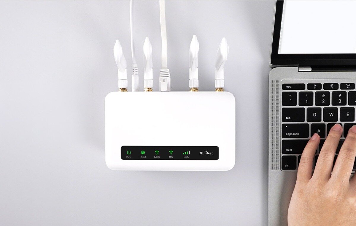 Widok z góry na biały router z czterema antenami, podłączony kablami. Obok stoi otwarty laptop, a na klawiaturze widać dłoń.