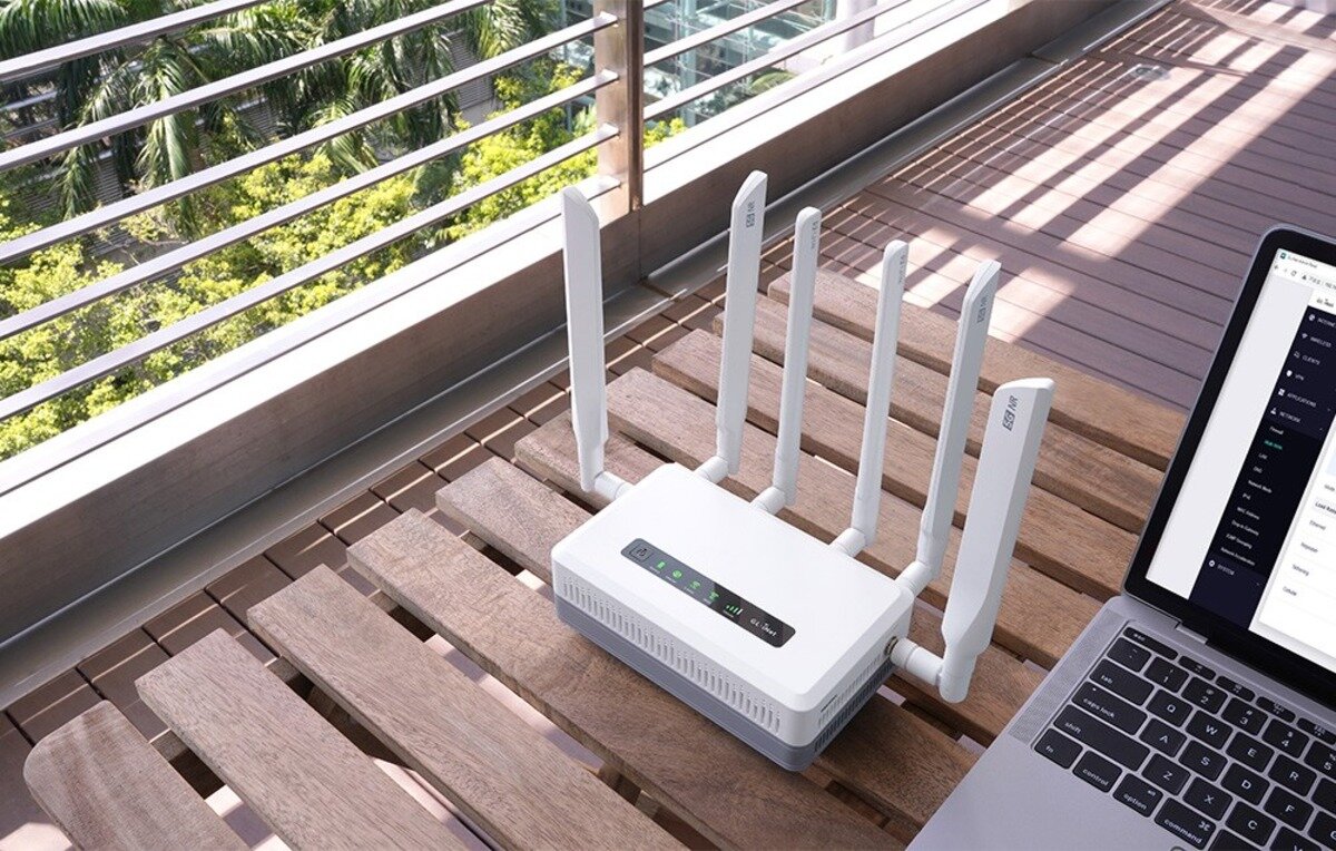 Biały router z sześcioma antenami stoi na drewnianym stoliku tarasowym obok otwartego laptopa