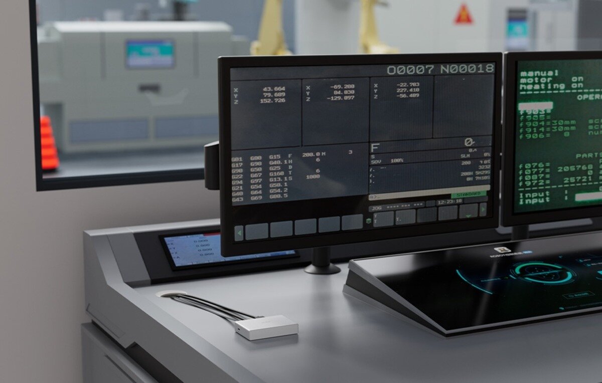 Na biurku znajdują się dwa monitory z wyświetlonymi danymi technicznymi oraz urządzenie przypominające przełącznik KVM. W tle widoczne jest wnętrze pomieszczenia z maszynami przemysłowymi.
