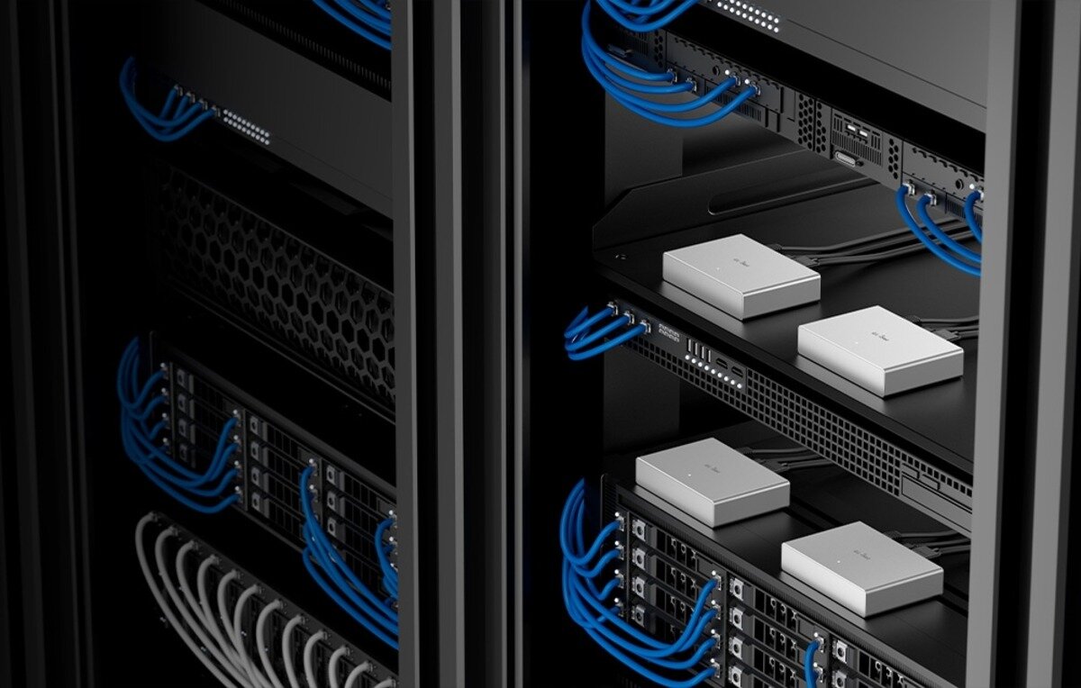 Obraz przedstawia wnętrze szafy serwerowej z kilkoma urządzeniami elektronicznymi i niebieskimi kablami sieciowymi. Na półkach znajdują się białe urządzenia o prostokątnym kształcie.