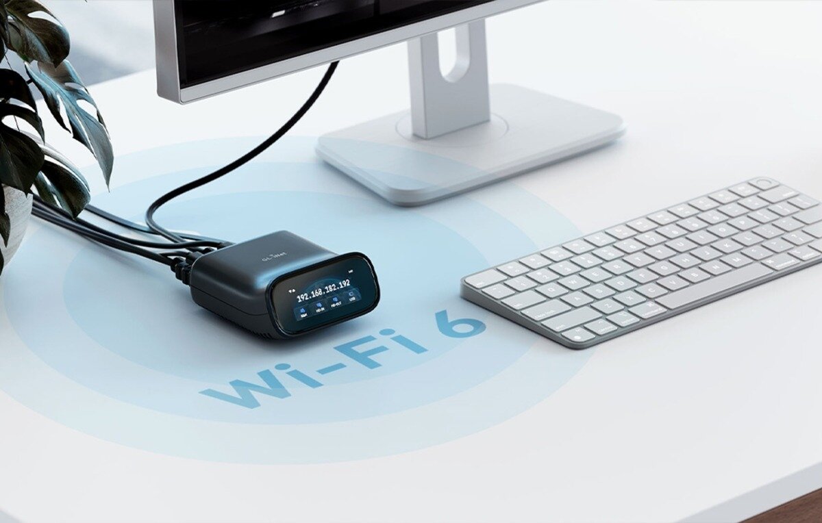 Na biurku znajduje się urządzenie z wyświetlaczem pokazującym adres IP, obok monitora i klawiatury. Na powierzchni biurka widoczny jest napis \'Wi-Fi 6\'.