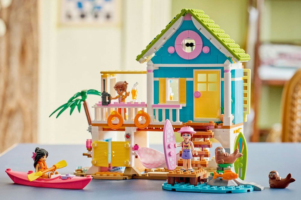 Zestaw LEGO przedstawia kolorowy domek na plaży z figurkami postaci i fokami. W tle widać elementy dekoracyjne, takie jak deska surfingowa i kajak.