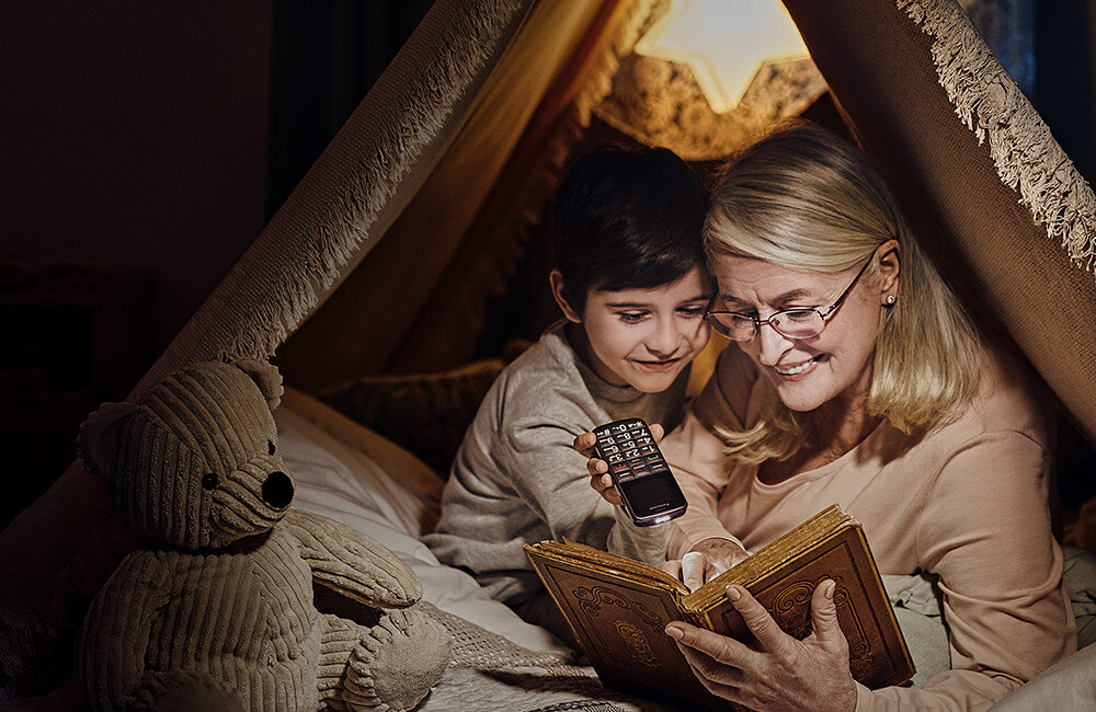 Dwie osoby siedzą w namiocie z koców, jedna z nich trzyma telefon z dużymi przyciskami. Obok znajduje się pluszowy miś, a w tle widać lampkę w kształcie gwiazdy.