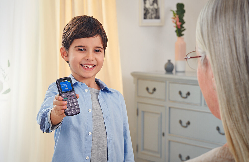 Dziecko trzyma czarny telefon z dużymi przyciskami i wyświetlaczem, na którym widoczny jest tekst. W tle znajduje się jasna komoda z dekoracjami.