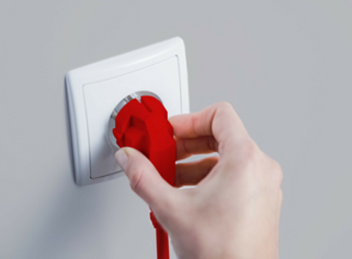 Osoba trzyma czerwoną wtyczkę, wkładając ją do białego gniazdka elektrycznego na szarej ścianie.