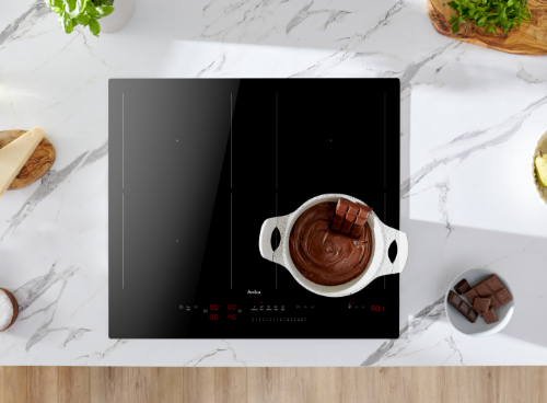 Na blacie kuchennym znajduje się czarna płyta indukcyjna z garnkiem pełnym czekolady i kawałkami czekolady na wierzchu. Obok płyty leży kostka czekolady na talerzyku, a na płycie widoczne jest logo \'Amica\'.