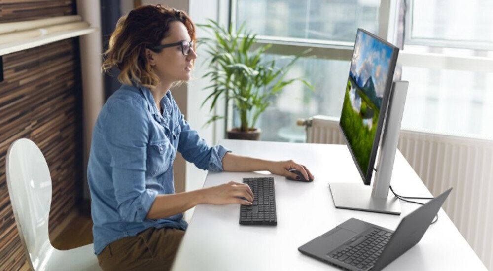 Osoba siedzi przy biurku, używając klawiatury i myszy przed dużym monitorem, na którym wyświetlany jest krajobraz. Obok monitora znajduje się otwarty laptop.