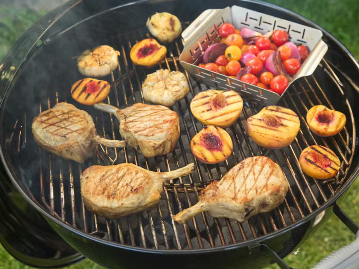 Na grillu węglowym znajdują się grillowane kotlety, połówki brzoskwiń oraz koszyk z pomidorkami koktajlowymi i cebulą. Grill jest okrągły, z metalowym rusztem.