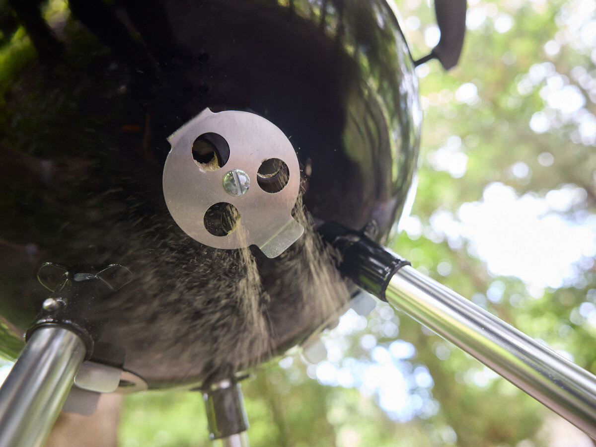 Zdjęcie przedstawia dolną część okrągłego grilla węglowego z metalowym regulatorem przepływu powietrza. Grill stoi na metalowych nogach, a w tle widać rozmyte drzewa.